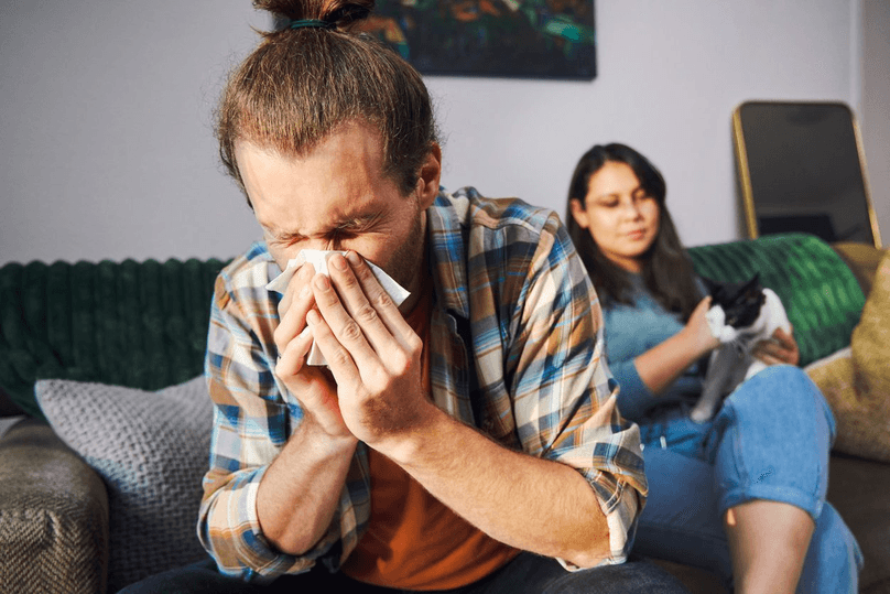 young man blowing his nose at home with his girlfriend and cat in background